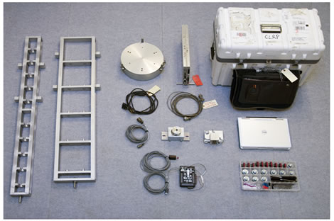 Figure 4. Photo. Calibration system hardware set. This photo shows the various components of a falling weight deflectometer (FWD) calibration system organized on a carpeted floor. The components include the geophone calibration stand, seismometer calibration stand, reference load cell, Vishay signal conditioner, ball-joint anchor, accelerometer box and calibration platter, Keithley KUSB data acquisition board with pushbutton, load cell and signal conditioner shipping case, FWD laptop with storage bag, geophone adapters, concrete anchors, universal serial bus key, handles for calibration stands, hex wrenches, and associated cables.