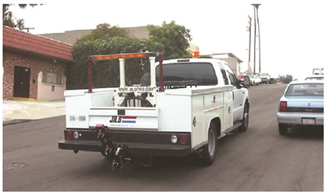 Figure 7. Photo. JILS-20T truck-mounted FWD. This photo shows a white JILS model 20T truck-mounted falling weight deflectometer (FWD) parked in the driving lane of an urban street. Cars are parked on both sides of the road where there is not a driveway.