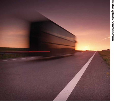 Photograph taken at dusk from beside a long, straight, two-lane highway, with white stripe heading off to the horizon and with a tractor trailer in motion blur speeding past in the left-hand lane.