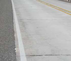 A short section of two-lane, jointed concrete pavement is shown close up with double yellow lines and an asphalt shoulder.