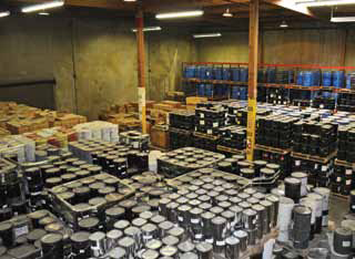 The interior of a warehouse is shown crowded with stacks of various containers, including tubes, cylinders, canisters, and barrels. The view includes hundreds of containers.