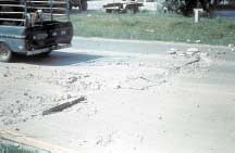 FIGURE 76.  Distress Type JCP 11 - A Blowup, Color photograph of jointed portland cement concrete pavement with distress type JCP 11 - blowup.  A high severity blowup with significant shattering of the concrete across two lanes is pictured.  There is upward movement at the pavement joint, especially at the line between the two lanes, and approximately 100-150 mm of the pavement joint depth is exposed due to loss of concrete in the inner wheel path near the center line joint.