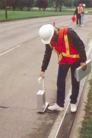 Figure B1.  The Georgia Faultmeter in use, Color photograph of the Georgia Faultmeter being used to measure faulting of jointed concrete pavement.  The photograph shows a transportation professional operating the Georgia Faultmeter at a transverse joint in a concrete pavement lane between the edge joint and the outer wheel path.  The base of the Faultmeter is set on the slab in the direction of traffic on the "leave side" of the joint.  The measuring probe contacts the slab on the approach side. 