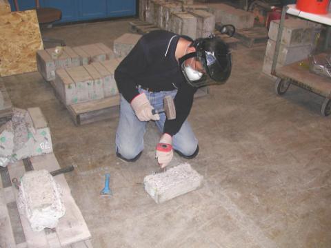 Figure 28. Extraction of embedded reinforcing bars. Photo. This picture shows a man indoors crouching on the floor. He is wearing protective kneepads, gloves, a mask and a face shield. He is using a hand-held sledgehammer to break apart the concrete to extract the reinforcing bars.