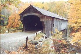 This long view shows the typical components that define a covered bridge: a timber structure supporting a deck surface that carries loads over an obstruction (usually a river). The structural components are protected from the elements by walls, roof and decks. This bridge is set on stone abutments with an unusual detailing of the portal and approach rail.