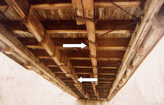 Existing bridge floors can have elements not part of the original construction. This picture shows two white arrows pointing to a twin line of longitudinal distribution beams that are aligned along the axis of the bridge and attached to the underside of the floor beams. Their intent is to distribute wheel load to more than one floor beam.