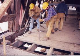 The picture shows two workers in hard hats prying up transverse nail-laminated decking with long crowbars. The vertical stringers and floor beams are exposed below.