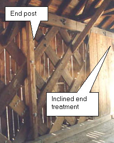 The picture shows the interior of the same bridge where the trusses require end posts back where the truss is still at full depth over the end support bearing points. The white text boxes point out the end post and the inclined end treatment at the edge of the picture. The interior of the overhang appears to be paneled with planks.