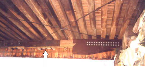 Another way to strengthen a covered bridge is to use supplemental steel rods, cables or rolled shapes to support existing members. The picture shows dark metal with holes over a bottom chord with the white arrow pointing to high-strength post-tensioning rods installed below and parallel to the chord.