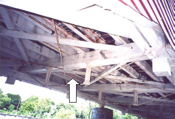 Floor beams can be modified by adding a steel assembly to post-tension the floor to duplicate an inverted queenpost truss. The picture shows the underside of a bridge with timber floor beams and wooden diagonals with the white arrow pointing to the reinforced steel assembly running diagonally across the floor deck. (Engineers will recognize this as a strutted beam.)