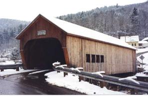 This diagonal view of the newly built bridge in winter shows snow on the roof and the banks, the extended portal with a sign, four windows, and metal side rails.