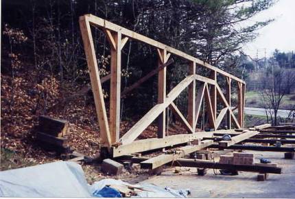 This picture shows the beginnings of floor members on blocks and the side wall with a queen post, diagonals,braces and wall framing, all propped up by timbers in the ground.