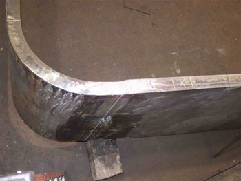 The photo shows a side view of field specimen FG40M-TF1-Curved-FCM with the weld oriented vertically. The specimen is comprised of curved plates of unequal thickness and equal width. The thicker plate is to the right in the photo.