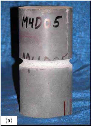 Photos. (a) Notched cylinder and (b) testing of an unnotched cylinder. (a) shows a 102-millimeter diameter cylinder after it has had a notch milled into its circumference at midheight. (b) shows an unnotched cylinder undergoing direct tension testing. This cylinder has been epoxied into the testing machine and an axial displacement measuring device has been attached to the specimen.
