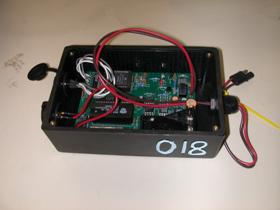 Figure 22. Photo. Data logger incorporating a ZRA. The figure shows an overhead photo of a data logger (black colored plastic box with 018 written on the right side in white) with the cover removed. The circuit board, which appears green, and the battery compartment are observed with connectors for the sensor (red- and black-paired wires) on the right and the data cable connector (black colored end cap) on the left end of data logger.