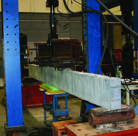 Figure 1. Photo. Test setup for cyclic loading of a beam. This figure shows the 15-inch (381 mm) deep ultra-high performance concrete (UHPC) beam in the load frame that was used to apply the flexural loads. The beam is supported by rollers at each end and is loaded at two points near midspan via rollers. A sodium chloride (NaCl)-soaked sponge is immediately under the constant moment region of the beam.