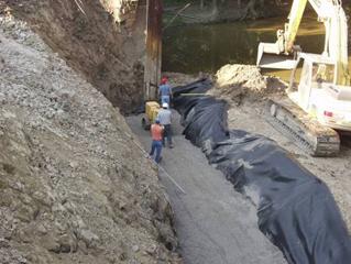 Photo showing the encapsulation of the fill in the reinforced soil foundation (RSF). The workers are compacting the crushed, well-graded gravel fill with a vibratory roller before folding the geotextile over the top of the RSF.