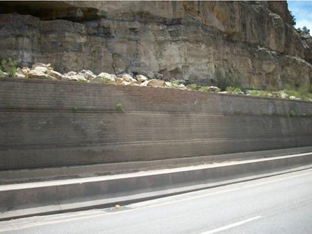 The photo shows the repair of the damaged geosynthetic reinforced soil (GRS) wall from figure 84. On the top of the wall is rubble from the breakdown of the boulders. The repaired patch is a slightly lighter color than the existing, undamaged GRS wall.