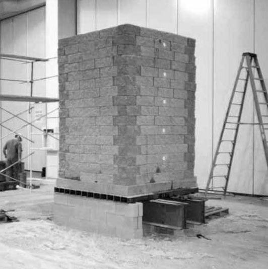 Photo showing the Vegas Mini-Pier Experiment. The geosynthetic reinforced soil (GRS) mass is on top of a concrete base, which is sitting on cinder blocks located at two edges. In the middle of the cinder blocks are steel beams. Scaffolding and a ladder are located behind the experiment.