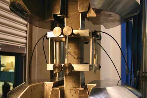 This photo shows a prismatic ultra-high performance concrete (UHPC) specimen gripped within a uniaxial tensile testing machine. The machine is gripping thin aluminum plates that have been epoxied to the surface of the UHPC prism. The gage length at the center of the UHPC specimen is encircled by a strain measuring device that measures the change in distance between the two planes at the ends of the gage length.