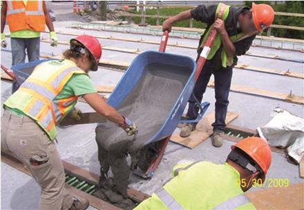 This photo shows fresh ultra-high performance concrete (UHPC) being poured from a wheelbarrow into a deck-level connection. The fluid UHPC flows into the connection around the non-contact lap-spliced reinforcing bars.