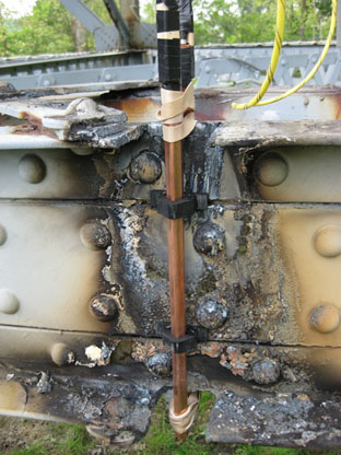 This photo is a close-up of a shape charge used in the explosive demolition. Running from left to right across the photo is the built-up chord member made from angles and plates riveted into a C-shape. In the center of the figure, the outstanding legs of the angles have been flame cut away for approximately 2 inches, flush to the web of the built-up C-shape. A copper clad rod (the shape charge) is clamped to the C-shape web in a vertical orientation where the outstanding legs had been removed.
