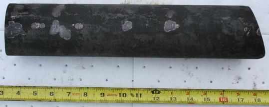 Figure 4. Photo. Received unthreaded sample after cleaning. This photo shows a bar of steel. A ruler in the foreground shows the overall length is approximately 15 inches. The width of the bar appears to be about 3 inches.