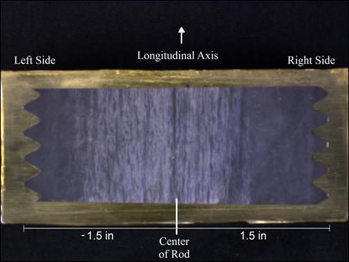 Figure 25. Photo. Mounted cross section of threaded rod etched with Marshall’s reagent. The photo shows a vertical section of a threaded anchor rod mounted in a yellow, semi-opaque epoxy. The mounted sample is placed on a black background. The longitudinal axis of the threaded rod is labeled with an upward vertical arrow. The left and right sides of the rod are labeled as is the center. Two scale bars begin at the center of the rod and end at the left and right edges of the threads, respectively. The scale bar on the left side is -1.5 inches, and the scale bar on the right is 1.5 inches. The rod has an approximate height of 1 inch, and the epoxy extends slightly past the rod on all four sides. The surface of the anchor rod is etched with Marshall’s reagent, which reveals light and dark areas of the microstructure. A region of approximately 0.5inches on the left side of the rod and approximately 0.75 inches on the right side of the rod are dark colored. The middle 1.75 inches of the rod shows alternating light and dark vertical bands.