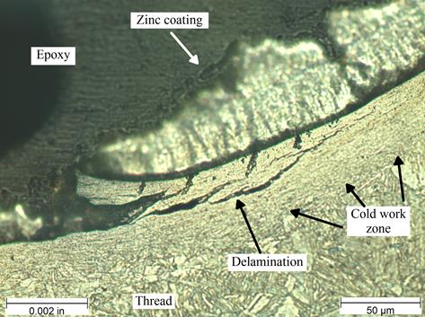 Figure 33. Photo. Micrograph of typical delamination at thread surface etched with 2‑percent nital at 500x magnification. This photo shows a micrograph that was taken of a thread surface from a sample mounted in epoxy and etched in 2-percent nital. It was viewed under bright field illumination at 500x magnification. A scale bar with a length of 0.002 inches is shown in the lower left corner of the photo, and a scale bar with a length of 50 micrometers is shown in the lower right corner. The epoxy mounting material appears black and is on the left side of the photo. The zinc coating can be seen on the thread surface. A region of cold work can be seen on the thread surface because of the compressed look of the microstructure. The image also shows a delamination that formed within the cold-worked region that follows the contour of the thread root.