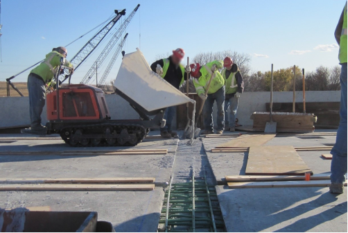 This photo shows the pour of a grout into the void space between prefabricated bridge elements (PBEs). The grout is pouring from a motorized buggy into an open strip between the reinforced concrete deck PBEs.