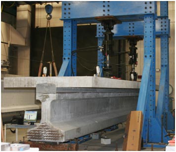 This photo shows an ultra-high performance concrete composite connection test specimen in the load frame during cyclic testing. The end of the prestressed girder/haunch/deck panel specimen is sitting on a roller support on the left side of the photo. The two load frame and actuators that apply vertical structural loading are straddling the simply supported specimen.
