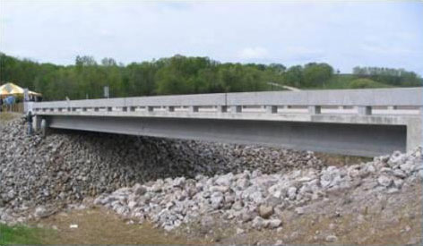 The photograph shows an oblique elevation view of the single-span bridge as viewed from the creek bank. The side of an exterior girder, the side of the bridge deck, and the side of the barrier railing is visible, along with the rip-rap protecting the stream bank from erosion.