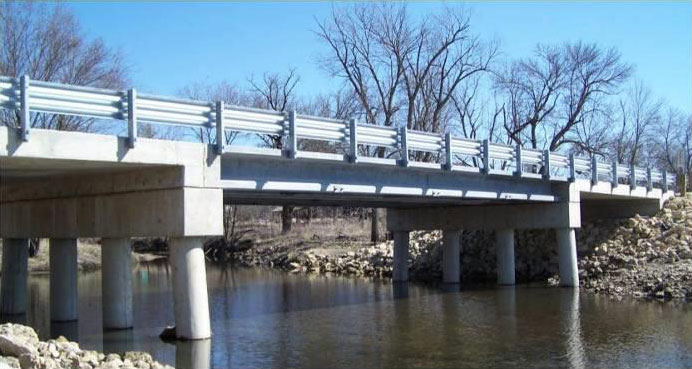 The photograph shows an oblique elevation view of the three-span bridge as viewed from the waterway bank. The sides of the reinforced concrete slab end spans as well as the ultra-high performance concrete pi-girder middle span are visible. Piers with pier caps are visible at the edges of the waterway.