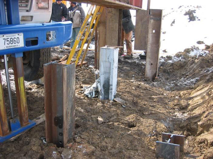 The photograph shows a precast ultra-high performance concrete pile after it has been driven into the ground. Three I-shaped steel piles that have also been driven into the ground are visible nearby.