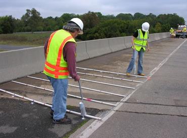 The photograph shows two workers on the shoulder of a bridge overpassing a highway in the foreground, and a utility truck on the bridge’s shoulder along with three additional workers in the rear of the photograph. The focus is on the activities of the two workers in the foreground. The workers are creating a data collection grid in order to accurately notate testing areas of the bridge. The workers are using six pieces of PVC to create a data collection grid – this is the jig. The jig has been placed with one end against the retaining wall and the other end touching the shoulder lane marker and the jigs are placed 2-feet apart, encompassing a 12-foot span of the bridge’s shoulder. Each jig has been marked with paint at 12-inch intervals, in alternating colors to assist the workers in the layout of the collection grid. The workers are using hand-held cans of water-soluble spray paint on an extension handle (so that bending is not required), to spray dots in a 2-foot by 2-foot grid pattern, using the black jig marking as a measurement guide.