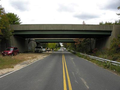 This photo displays a view of the New Jersey pilot bridge in Upper Freehold, NJ, as shown from Sharon Station Road. The bridge spans horizontally across the image. There are four inspectors underside of the bridge. Three inspectors are on the ground conducting visual inspection. One inspector is using a van with a lift, to perform an in-depth visual inspection to the girders.