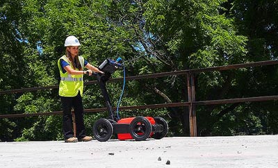 In this figure, a worker is pushing a ground penetrating radar unit on the deck of a bridge. The unit has two wheels in front and two in the rear. A red plastic box between the wheels is in contact with the deck surface. Above the red box is a rectangular control unit resting at an angle on the framework extending from the wheels to the worker’s hands. A computer is at the top of the unit, almost at the worker’s eye level.
