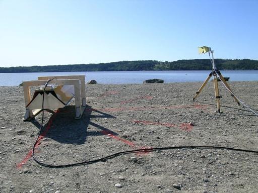 Figure 29. Photo. Photograph of outdoor test configuration, including B1823 SF GPR antenna and horn measurement antenna (side view). The SF GPR antenna is suspended above the ground by two white saw horses. The ground surface is gravel. A lake is visible in the background. The picture is taken from the side of the measurement antenna and the SF GPR antenna. The side of the measurement antenna is visible in the right of the picture while the end of the SF GPR antenna suspended from the two saw horses is visible on the left of the picture.