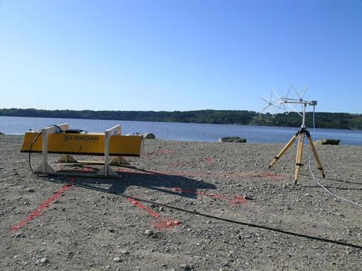Figure 30. Photo. Photograph of outdoor test configuration, including B1823 SF GPR antenna with its long axis in line with the measurement direction of the white biconic measurement antenna. The SF GPR antenna is suspended above the ground by two white saw horses. The ground surface is gravel. A lake is visible in the background. The picture is taken from the side of the measurement antenna and the SF GPR antenna.
