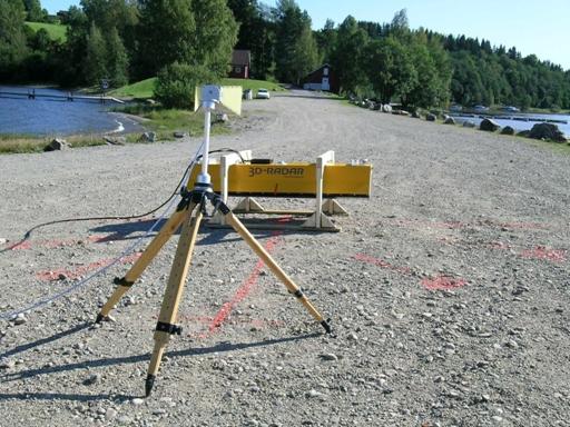Figure 31. Photo. Photograph of outdoor test configuration, including the yellow trapezoidal B1823 SF GPR antenna and white horn measurement antenna sitting about four feet above the ground on a three legged yellow tripod (boresight view). The SF GPR antenna is behind the measurement antenna and resting perpendicular to its line of sight. The SF GPR antenna is suspended above the ground by two white saw horses. The ground surface is gravel. A lake is visible in the background. The picture is taken from behind the measurement antenna with the SF GPR antenna on the far side of the measurement antenna.