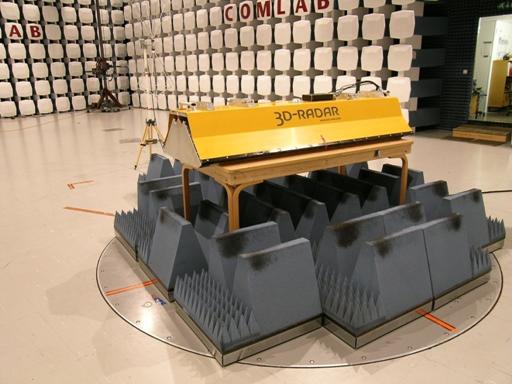 Figure 32. Photo. Photograph of indoor test configuration, including anechoic chamber. The yellow trapezoidal SF GPR unit is resting on a set of grey blocks sitting on a beige floor. The walls are patterned with white square emissions absorbing blocks. 
