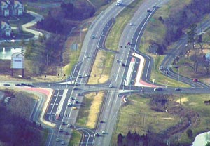 Aerial photograph of a displaced left-turn intersection in which traffic wishing to turn left from a divided highway onto a cross-street diverges via dedicated left turn lanes that cross opposing traffic in advance of the intersection, then merge into the cross-street traffic.