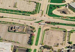 Aerial photograph of a restricted crossing U-turn intersection in which a minor road intersects a major six-lane road. At the intersection, the through movements for the minor road are blocked by a median, although left-turn movements are allowed from the major road to the minor roads. Openings in the median are provided on either side of the intersection on the major road that allow vehicles to make a U-turn. In this configuration, the through traffic from the minor street must make a right turn into the main street, make a U-turn in the median, and take a right turn to continue on the minor road.