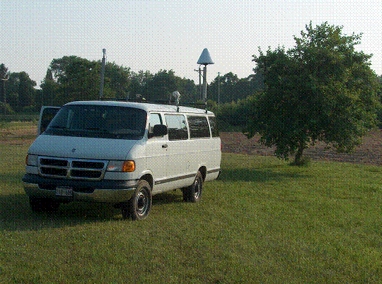 Figure 25. Photo. Van configuration for multipath testing at the Hagerstown GWEN site for the NDGPS 700829 (3) antenna from Ashtech to van 700829 (3) antenna from Ashtech test. Picture of a large white van with two antennas mounted on top.  The van is facing toward the lower left hand corner of the photo. On the left side of the van toward the front is a HA-NDGPS antenna; on the right side of the van is mounted the GPS antenna.