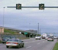 This photograph shows a several cars traveling on a roadway. Two illuminated signs hang above the right and middle lanes. The sign over the right lane shows that cars in that lane can only make a right turn. The sign over the middle lane indicates that cars in that lane can make a right turn or continue traveling straight.