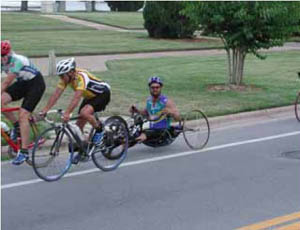 Figure 49: Photo. A hand cyclist. A man is riding a hand cycle in a bicycle lane. Two other bicyclists ride just in front of him.
