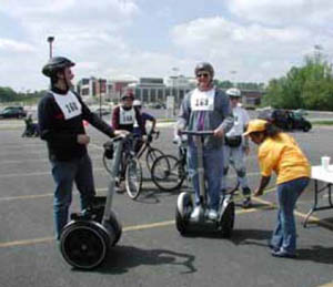 Figure 50: Photo. Segway users at the physical measurements station. An event staff person is taking measurements of a participant standing on a Segway. Another participant beside a Segway, a participant using inline skates, and two participants on bicycles are waiting to be measured.