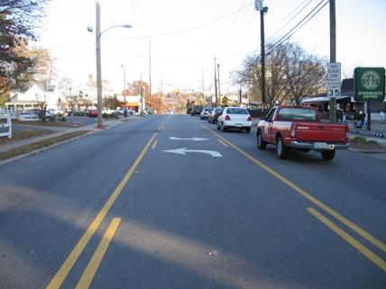 Figure 1. Photo. Example of a TWLTL in North Carolina. This photo shows a road divided into three lanes where the center lane is a turn lane, designated by solid yellow lines to the outside of each side and broken yellow lines on the inside of the lane. Within the lane, left-turn arrows are marked from each direction on the road. The road is in a suburban area, with small businesses and residential buildings on either side. The speed limit is 56.35 km/h (35 mi/h), and there is a traffic light toward the background of the photo. There is traffic in both lanes of the road.