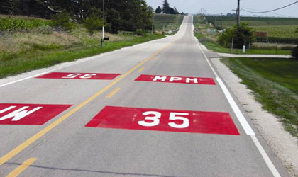 Photograph showing a two-lane highway with red patches painted on the pavement with the number "35" painted white in the center of one red patch and the letters "MPH" painted  white in a second red patch immediately downstream. The red patches with the "35"  and "MPH" legends are painted in both directions of travel. Because the red  background markings appear freshly painted, the white "35 MPH" legends really stand out in the photograph.