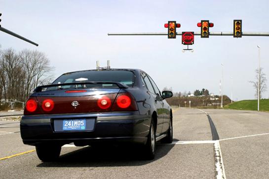 This picture shows a car stopped at the stop line of the intersection on the Virginia Smart Road. The mast arm warning that is shown in figure 6 can be seen at the far side of the intersection. In the photo, the road curves gently to the left at the intersection.